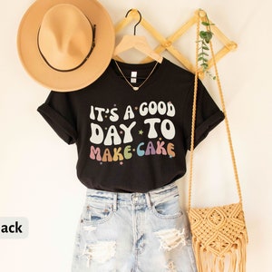 Peut inclure: T-shirt noire avec le texte "It's a good day to make cake" en couleurs arc-en-ciel avec des &eacute;toiles. Le t-shirt est suspendu &agrave; un portant en bois avec un sac en paille et un chapeau.