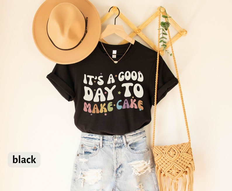 Peut inclure: T-shirt noire avec le texte "It's a good day to make cake" en couleurs arc-en-ciel avec des &eacute;toiles. Le t-shirt est suspendu &agrave; un portant en bois avec un sac en paille et un chapeau.