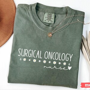 May include: A green t-shirt with the text "SURGICAL ONCOLOGY nurse" in white lettering with a small heart and daisies. The shirt is made by Comfort Colors.