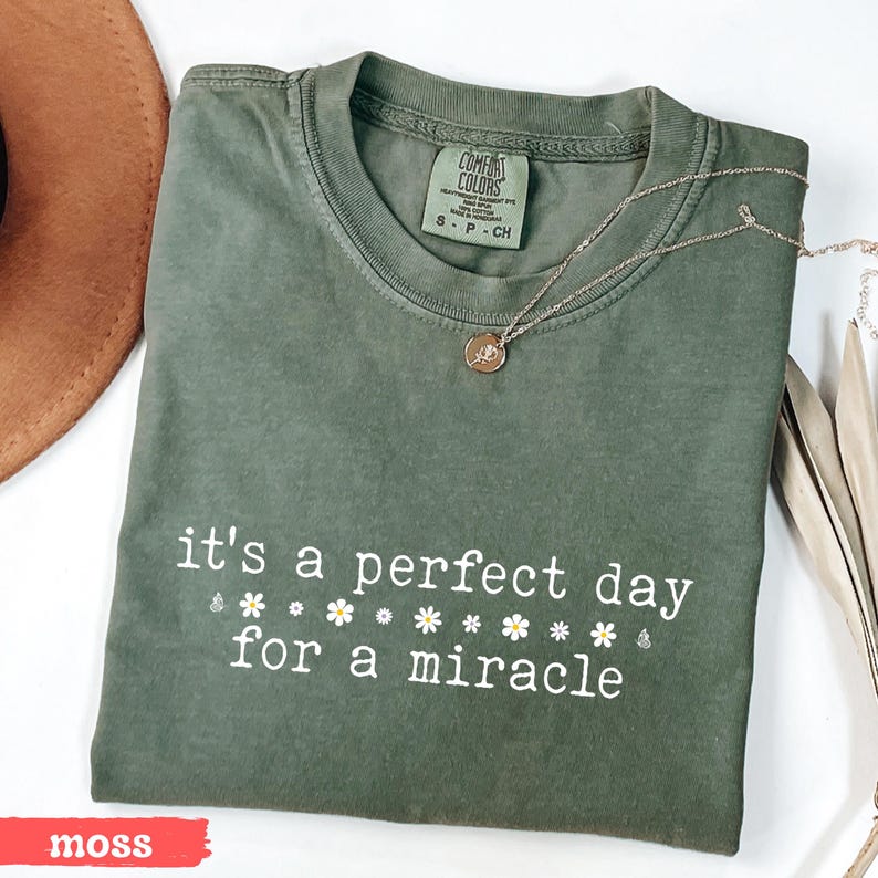 May include: A moss green t-shirt with the text "it's a perfect day for a miracle" in white lettering with small white daisy flowers. The shirt is made of a soft, comfortable material.