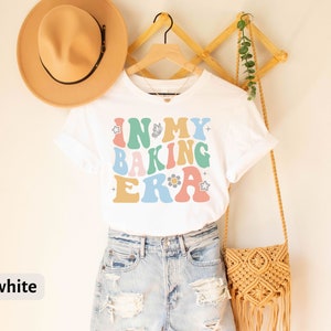 May include: White t-shirt with the text "In My Baking Era" in colorful retro-style lettering. The shirt is being modeled on a person wearing denim shorts and a straw hat.