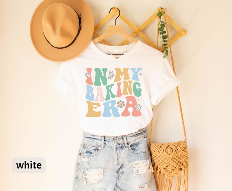 May include: White t-shirt with the text "In My Baking Era" in colorful retro-style lettering. The shirt is being modeled on a person wearing denim shorts and a straw hat.