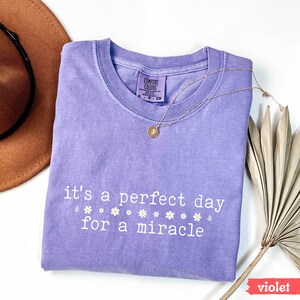 May include: A purple t-shirt with the text "it's a perfect day for a miracle" and a row of white daisies printed on the front. The shirt is folded and laid flat on a white surface.