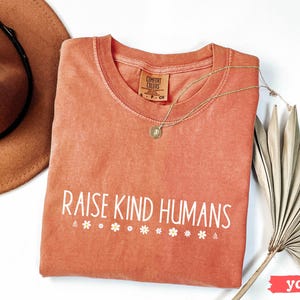 Op de afbeelding: Een perzikkleurig T-shirt met de tekst "Raise Kind Humans" in witte letters. Het shirt heeft een verweerde look en is versierd met kleine witte bloemenaccenten.