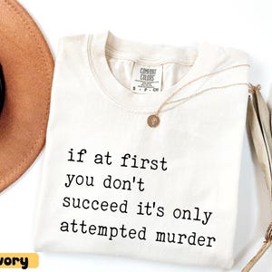 Op de afbeelding: Ivoor Comfort Colors T-shirt met de zwarte tekst "if at first you don't succeed it's only attempted murder". Een gouden ketting met hanger ligt op het shirt. Een bruine hoed is zichtbaar op de achtergrond.
