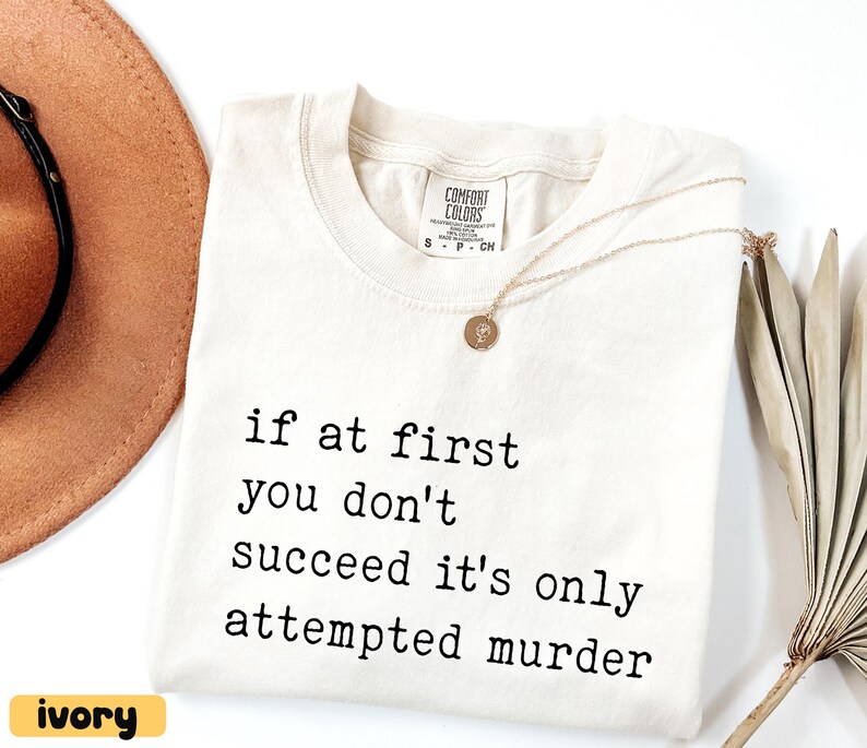 Op de afbeelding: Ivoor Comfort Colors T-shirt met de zwarte tekst "if at first you don't succeed it's only attempted murder". Een gouden ketting met hanger ligt op het shirt. Een bruine hoed is zichtbaar op de achtergrond.