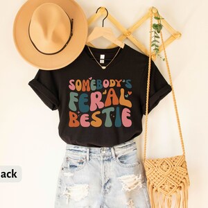 May include: Black t-shirt with the text "Somebody's Feral Bestie" in colorful, retro-style lettering. The shirt is worn with blue denim shorts and a crocheted bag.