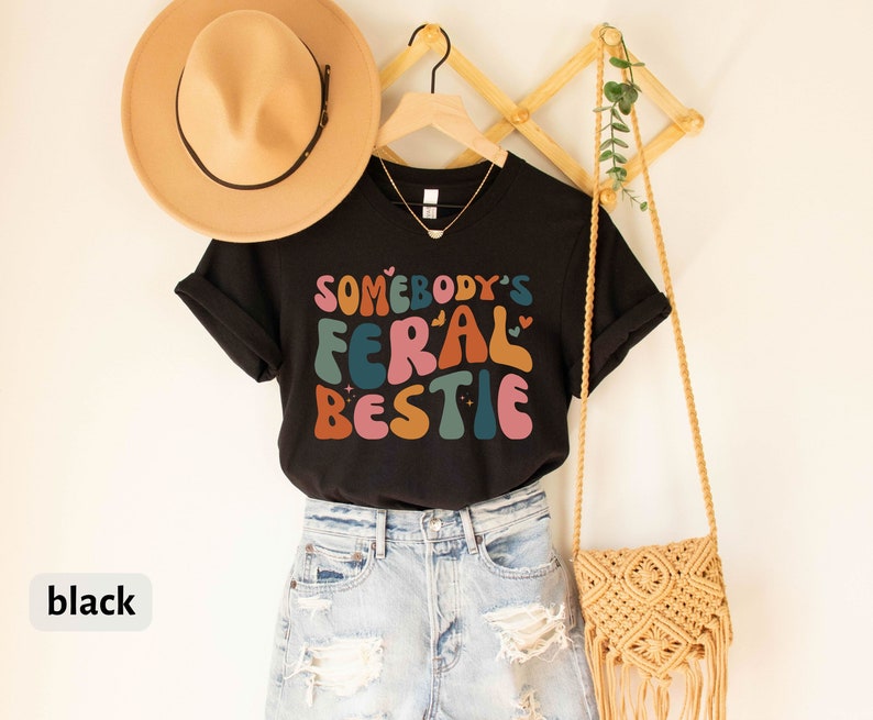 May include: Black t-shirt with the text "Somebody's Feral Bestie" in colorful, retro-style lettering. The shirt is worn with blue denim shorts and a crocheted bag.