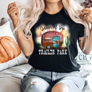 May include: A black t-shirt with the text "Queen of the Trailer Park" and a cartoon image of a pink camper with a turquoise door and brown wheels. The camper is set against a desert landscape with cacti and a sunset.