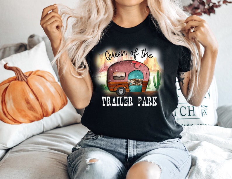 May include: A black t-shirt with the text "Queen of the Trailer Park" and a cartoon image of a pink camper with a turquoise door and brown wheels. The camper is set against a desert landscape with cacti and a sunset.