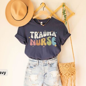 May include: A navy blue t-shirt with the text "Trauma Nurse" in rainbow colored letters with stars. The shirt is on a hanger and is being modeled with a straw hat and a crocheted bag.