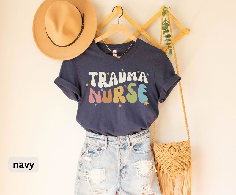 May include: A navy blue t-shirt with the text "Trauma Nurse" in rainbow colored letters with stars. The shirt is on a hanger and is being modeled with a straw hat and a crocheted bag.