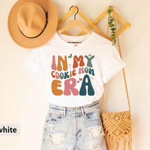 May include: White t-shirt with a colorful graphic that says "In My Cookie Mom Era".