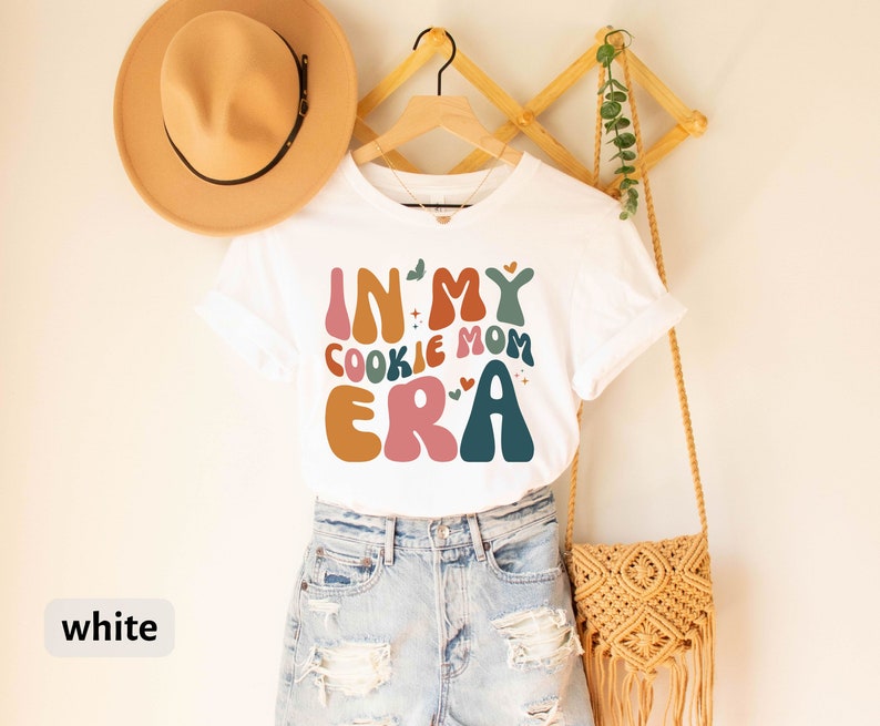 May include: White t-shirt with a colorful graphic that says "In My Cookie Mom Era".