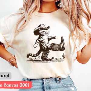 May include: Natural-colored t-shirt featuring a black and white illustration of a cat wearing a cowboy hat and boots. The cat is walking with a confident stride. The text "natural" is at the bottom left.