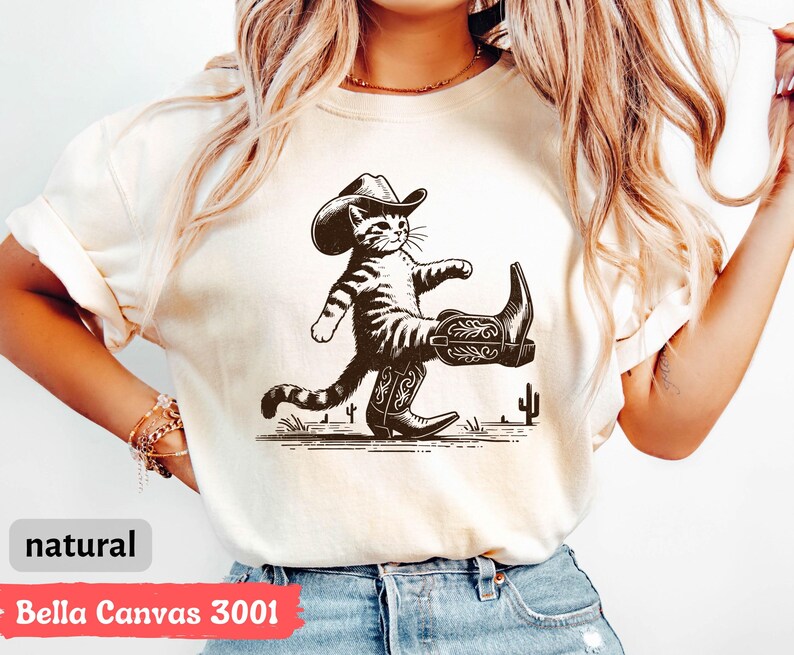 May include: Natural-colored t-shirt featuring a black and white illustration of a cat wearing a cowboy hat and boots. The cat is walking with a confident stride. The text "natural" is at the bottom left.