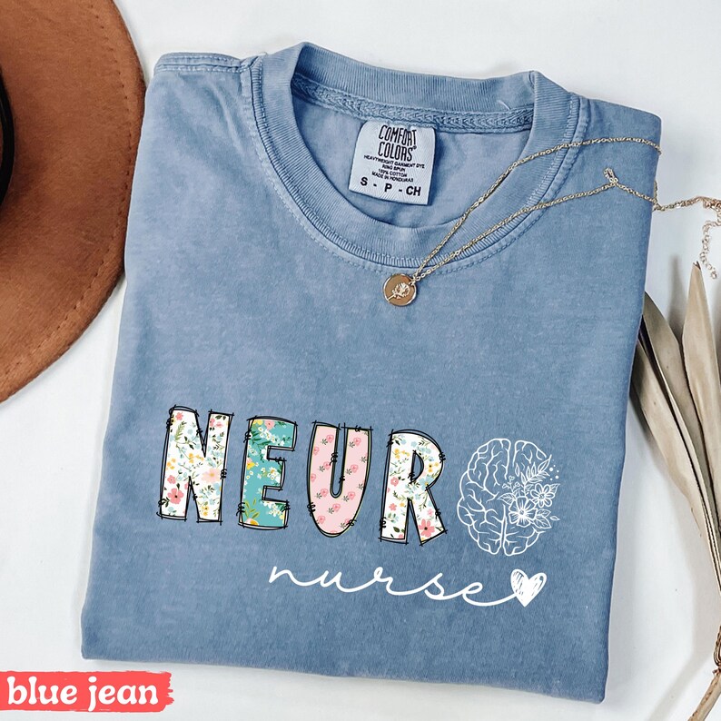 Puede incluir: Camiseta azul vaquero con la palabra "NEURO" en estampado floral y un gr&aacute;fico de cerebro. La palabra "nurse" est&aacute; escrita en cursiva debajo del gr&aacute;fico. Un collar dorado con un colgante est&aacute; sobre la camiseta.