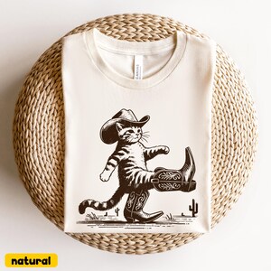 May include: Cream-colored t-shirt featuring a cartoon cat wearing a cowboy hat and boots, walking with one leg raised. The design is in brown tones, with a cactus in the background. The word "natural" is in a yellow rectangle at the bottom.