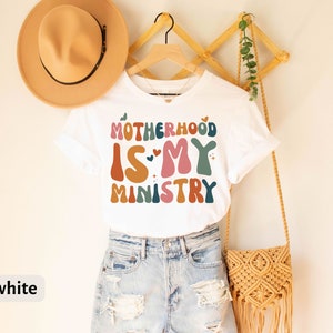 May include: White t-shirt with the text "Motherhood is my ministry" in colorful, retro-style lettering.