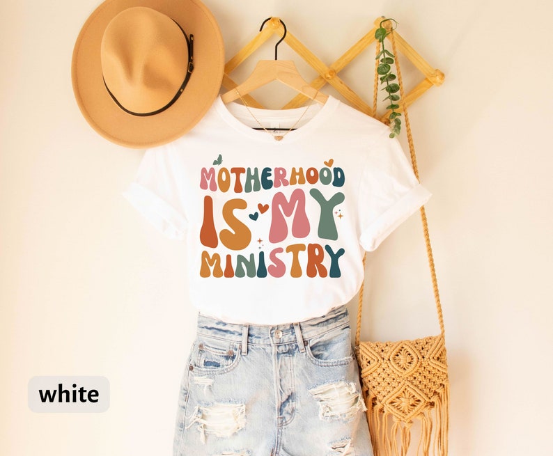 May include: White t-shirt with the text "Motherhood is my ministry" in colorful, retro-style lettering.