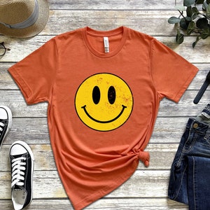 May include: A burnt orange t-shirt featuring a yellow smiley face graphic. The smiley face has black eyes and a black mouth.