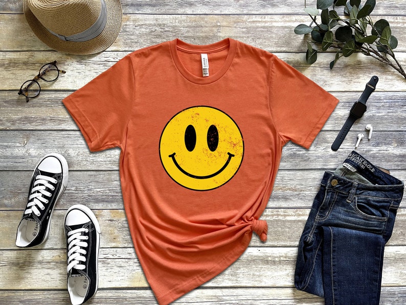 May include: A burnt orange t-shirt featuring a yellow smiley face graphic. The smiley face has black eyes and a black mouth.