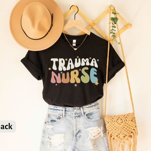 May include: Black t-shirt with the text "Trauma Nurse" in colorful, retro-style lettering with stars. The shirt is hanging on a wooden rack with a straw hat and a woven bag.