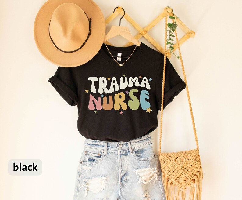 May include: Black t-shirt with the text "Trauma Nurse" in colorful, retro-style lettering with stars. The shirt is hanging on a wooden rack with a straw hat and a woven bag.