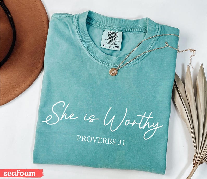 May include: Seafoam green t-shirt with the words "She is Worthy" and "Proverbs 31" in white script. A gold necklace with a pendant rests on the shirt. The shirt is folded, and a brown hat is partially visible.