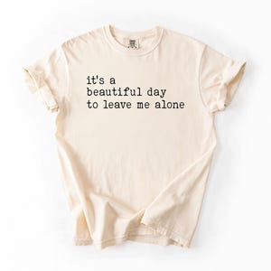 May include: A beige t-shirt with the text "it's a beautiful day to leave me alone" printed in black.