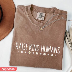 Op de afbeelding: Een bruin T-shirt met de tekst "Raise Kind Humans" in witte letters en een rij witte madeliefjes onder de tekst. Het shirt ligt op een wit oppervlak. Het woord "espresso" staat in rood onderaan de afbeelding.
