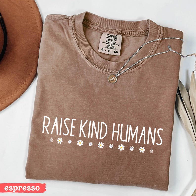 Op de afbeelding: Een bruin T-shirt met de tekst "Raise Kind Humans" in witte letters en een rij witte madeliefjes onder de tekst. Het shirt ligt op een wit oppervlak. Het woord "espresso" staat in rood onderaan de afbeelding.
