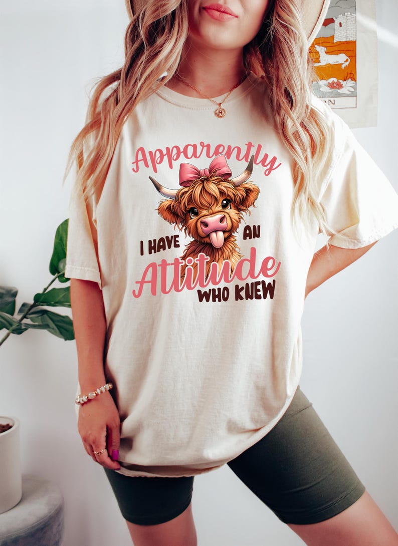 May include: A white t-shirt with a cartoon highland cow wearing a pink bow. The text on the shirt reads "Apparently I have an attitude who knew".