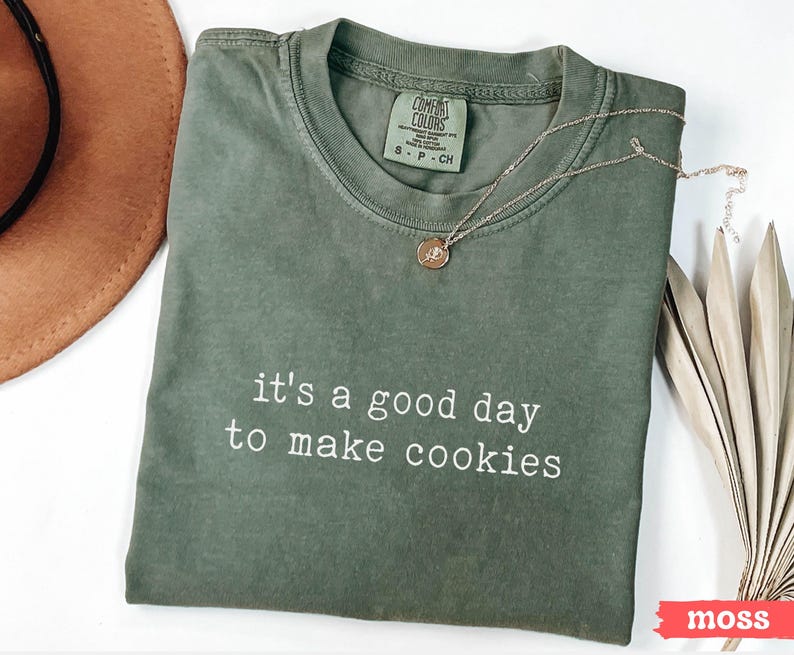 May include: A green t-shirt with the text "it's a good day to make cookies" in white lettering. The shirt is folded and laid flat on a white surface.
