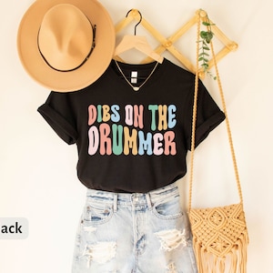 Puede incluir: Camiseta negra con el texto "Dibs on the Drummer" en letras de colores. La camiseta est&aacute; colgada en un perchero de madera con un sombrero de paja y un bolso tejido.