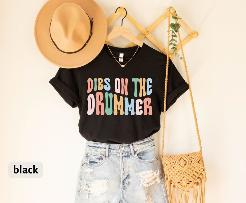 Puede incluir: Camiseta negra con el texto "Dibs on the Drummer" en letras de colores. La camiseta est&aacute; colgada en un perchero de madera con un sombrero de paja y un bolso tejido.