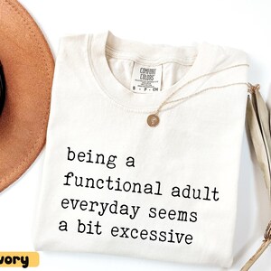 K&ouml;nnte beinhalten: Elfenbeinfarbenes T-Shirt mit dem Text "being a functional adult everyday seems a bit excessive" in Schwarz. Eine goldene Halskette mit Anh&auml;nger liegt auf dem Shirt. Ein brauner Hut und eine getrocknete Pflanze sind im Hintergrund.