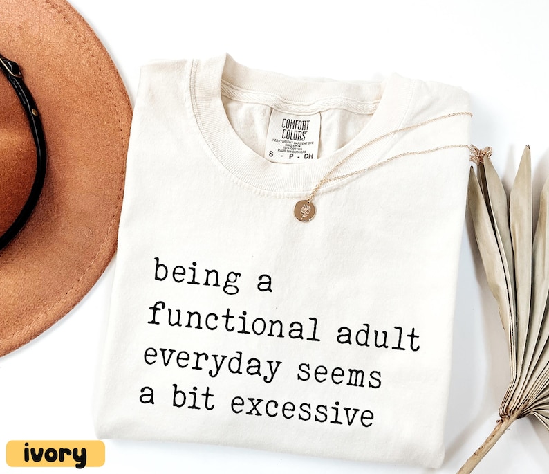 K&ouml;nnte beinhalten: Elfenbeinfarbenes T-Shirt mit dem Text "being a functional adult everyday seems a bit excessive" in Schwarz. Eine goldene Halskette mit Anh&auml;nger liegt auf dem Shirt. Ein brauner Hut und eine getrocknete Pflanze sind im Hintergrund.