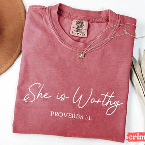 May include: Crimson-colored t-shirt with the words "She is Worthy" and "Proverbs 31" in white script. A silver necklace with a pendant rests on the shirt. The shirt is a Comfort Colors brand.