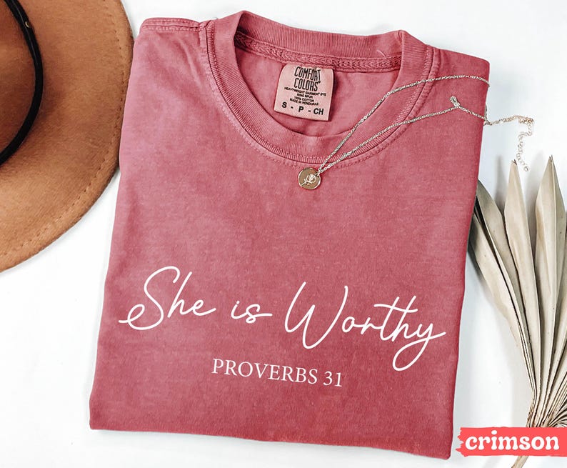 May include: Crimson-colored t-shirt with the words "She is Worthy" and "Proverbs 31" in white script. A silver necklace with a pendant rests on the shirt. The shirt is a Comfort Colors brand.