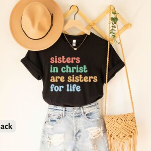 May include: Black t-shirt with the text "sisters in christ are sisters for life" in rainbow colors. The shirt is hanging on a wooden hanger and is styled with a straw hat and a woven bag.