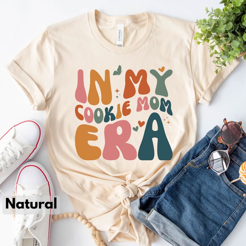 May include: A beige t-shirt with the text "In My Cookie Mom Era" in colorful, retro-style lettering. The shirt has a relaxed fit and a crew neck.
