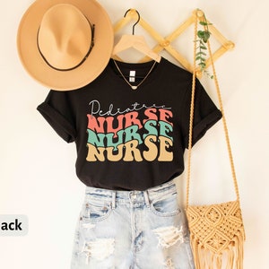 K&ouml;nnte beinhalten: Schwarzes T-Shirt mit dem Text "Pediatric Nurse" in einer Retro-Schrift mit bunten Buchstaben. Das Shirt ist auf einem Kleiderb&uuml;gel mit einem Strohhut und einer geflochtenen Tasche abgebildet.