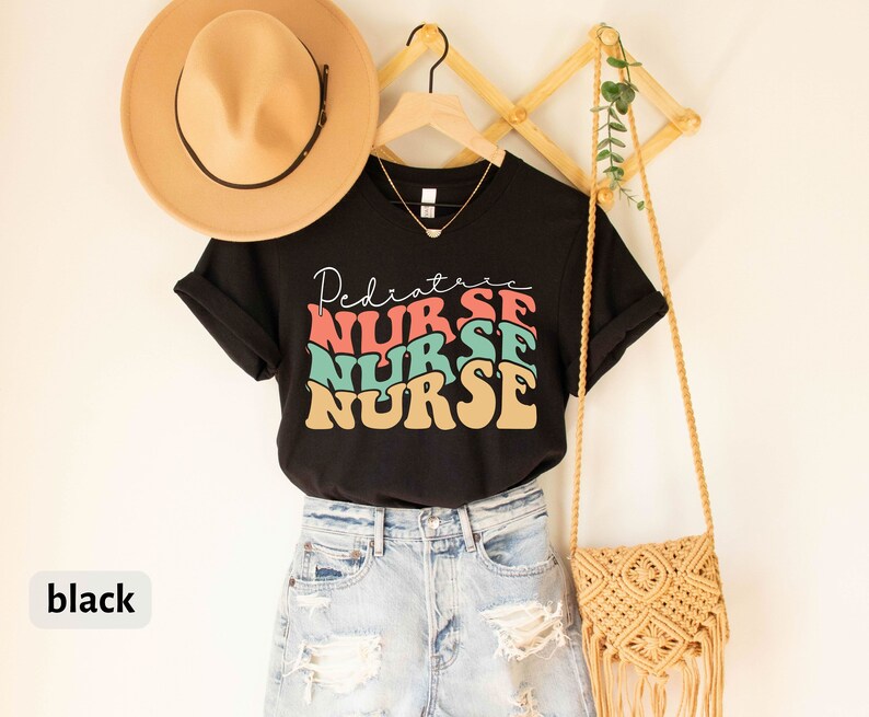 K&ouml;nnte beinhalten: Schwarzes T-Shirt mit dem Text "Pediatric Nurse" in einer Retro-Schrift mit bunten Buchstaben. Das Shirt ist auf einem Kleiderb&uuml;gel mit einem Strohhut und einer geflochtenen Tasche abgebildet.