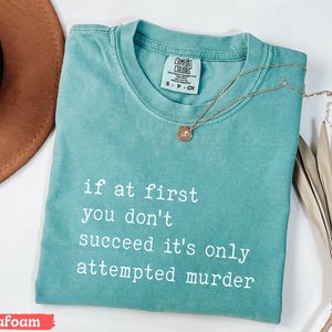 Op de afbeelding: Zeegroen t-shirt met de witte tekst "if at first you don't succeed it's only attempted murder". Een gouden ketting met een hanger ligt op het shirt. Het shirt is opgevouwen en heeft een Comfort Colors label.