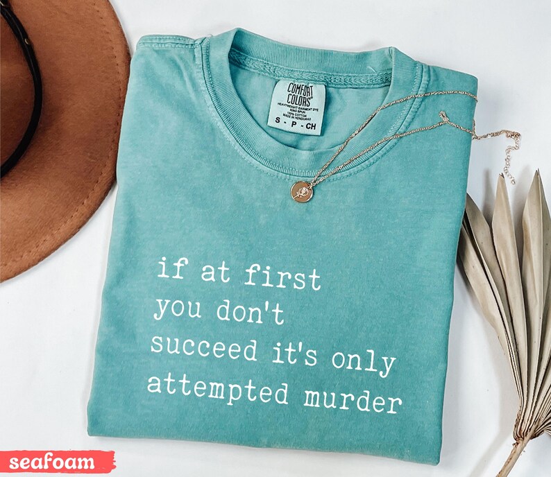 Op de afbeelding: Zeegroen t-shirt met de witte tekst "if at first you don't succeed it's only attempted murder". Een gouden ketting met een hanger ligt op het shirt. Het shirt is opgevouwen en heeft een Comfort Colors label.