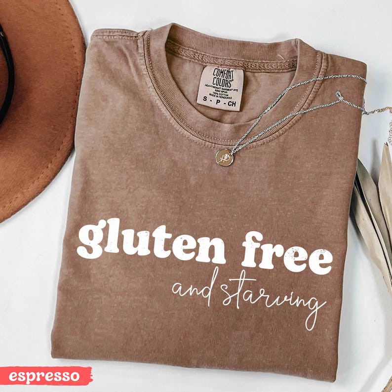 Puede incluir: Camiseta color espresso con las palabras "gluten free and starving" en escritura blanca. Un collar plateado con un peque&ntilde;o colgante descansa sobre la camiseta. La camiseta es de la marca Comfort Colors.