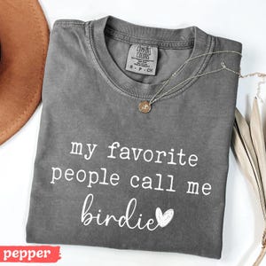 May include: A grey Comfort Colors t-shirt with the text "my favourite people call me birdie" in white. A gold necklace with a pendant is on the shirt. A brown hat is in the background.