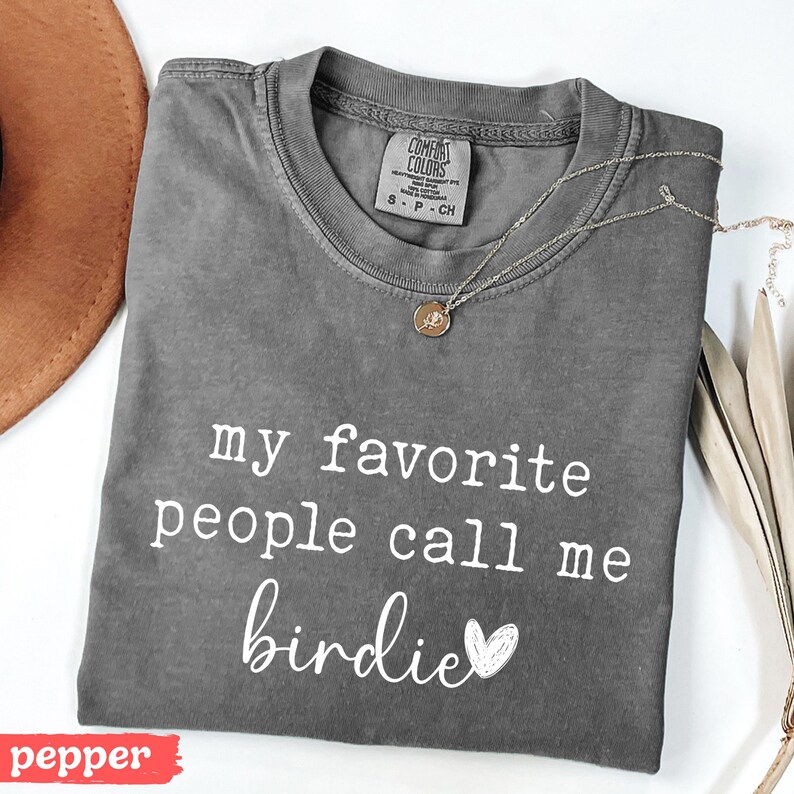 May include: A grey Comfort Colors t-shirt with the text "my favourite people call me birdie" in white. A gold necklace with a pendant is on the shirt. A brown hat is in the background.