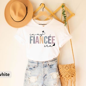 May include: White t-shirt with the text "in my fiancee era" in colorful letters with a ring graphic above the word "fiancee".